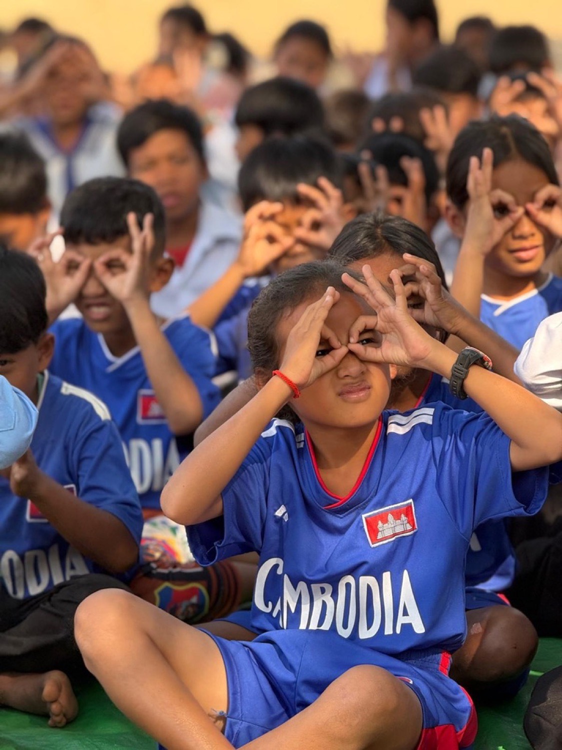 Children in blue Cambodia jerseys making circles with their hands over their eyes during a game