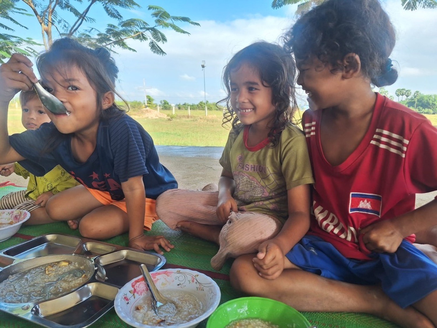 Children sitting together on a mat outdoors, eating and laughing
